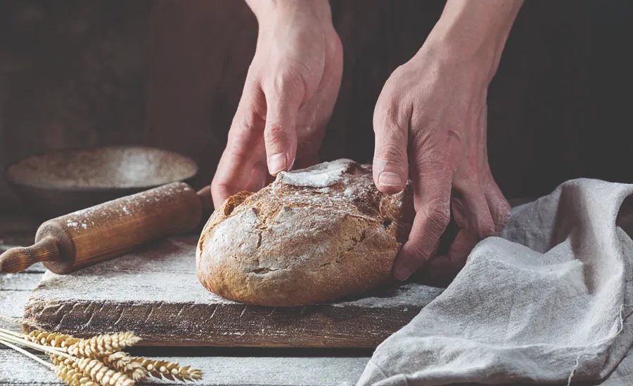 Hender skjærer opp ferskt brød på kjøkkenbenk med mel og bakeutstyr